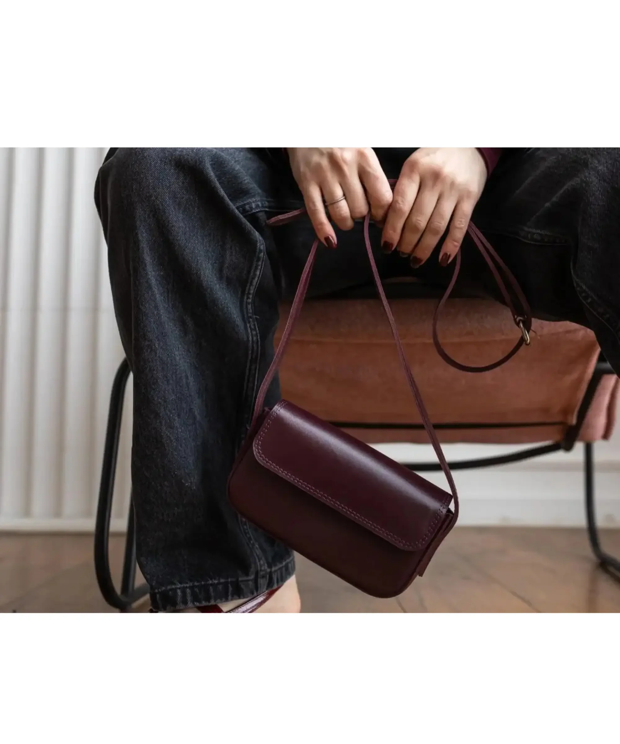 Burgundy genuine leather mini crossbody bag with a slim strap, held by a woman wearing dark jeans while seated on a chair, showcasing a minimalist and elegant design.