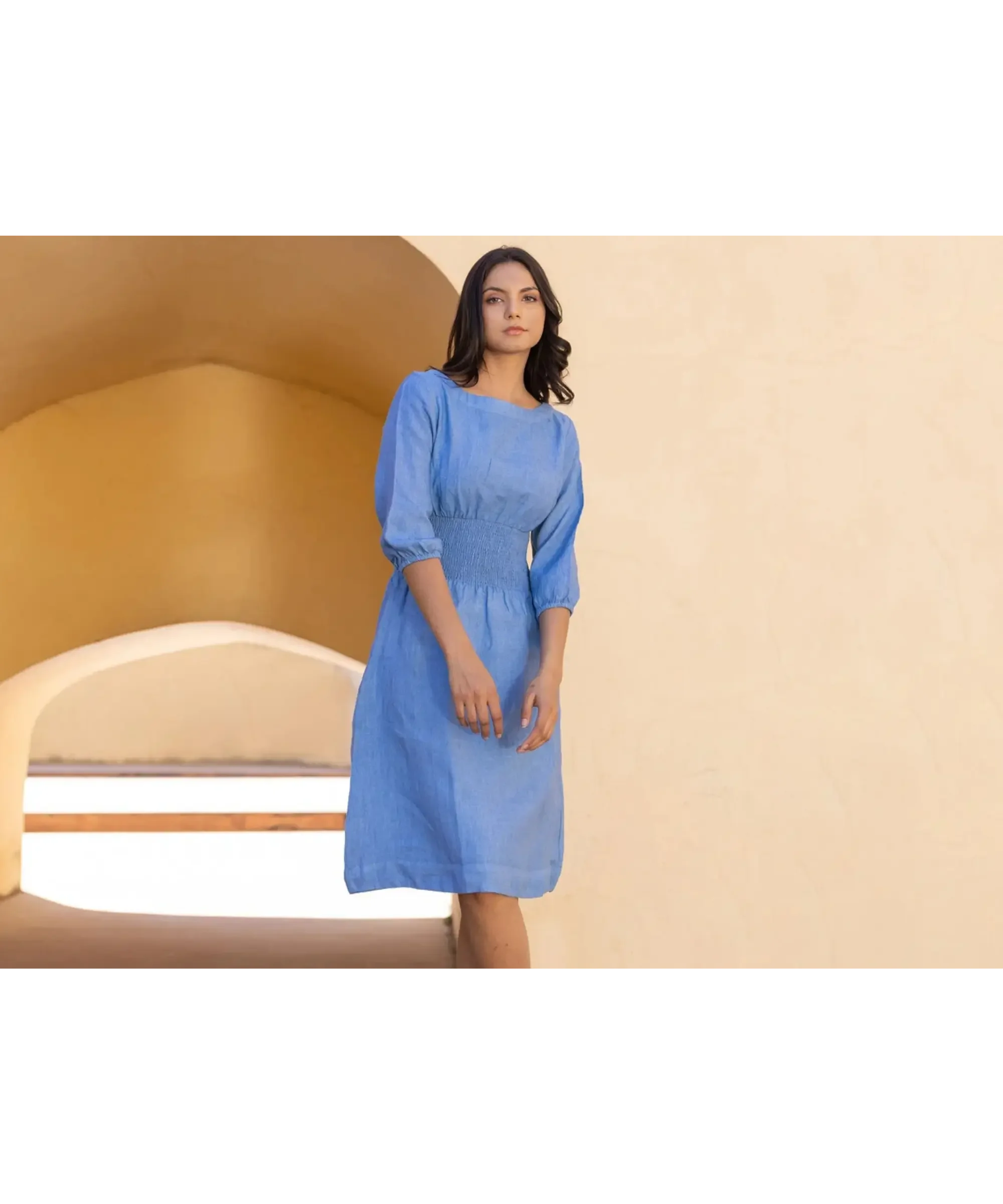 Blue linen midi dress with a softly ruched waist and three-quarter sleeves, featuring a relaxed silhouette and practical pockets, worn by a woman in a warm, minimalist architectural setting.