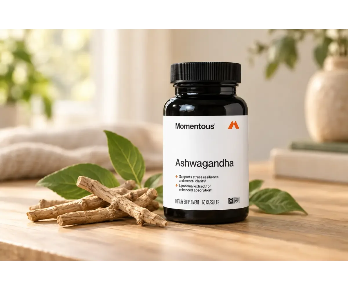 Momentous Ashwagandha supplement bottle displayed beside ashwagandha roots and green leaves on a softly lit wooden table.