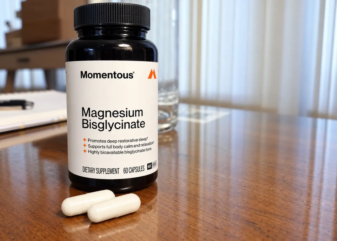 Momentous Magnesium Bisglycinate bottle with two white capsules on a wooden table, with a notebook and glass water bottle in the background.