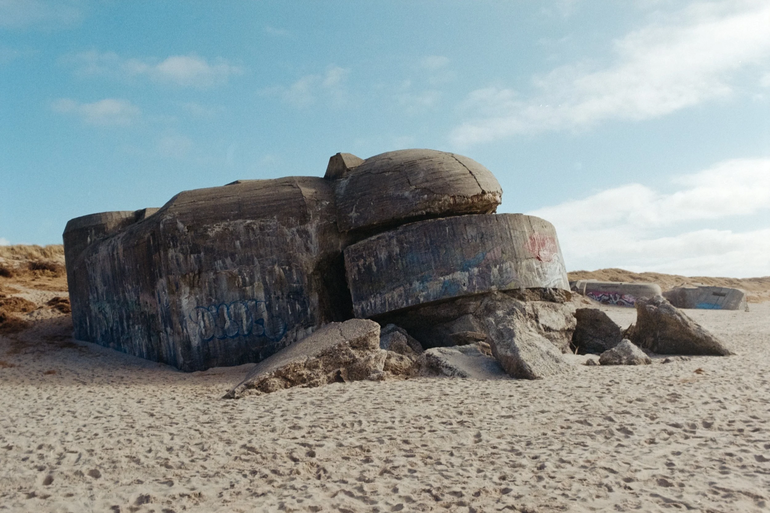 Analoge Fotografie von großer Bunkeranlage am Strand von Søndervig, Dänemark.
Im Hintergrund zieht sich eine Dünenlandschaft den Horizont entlang.