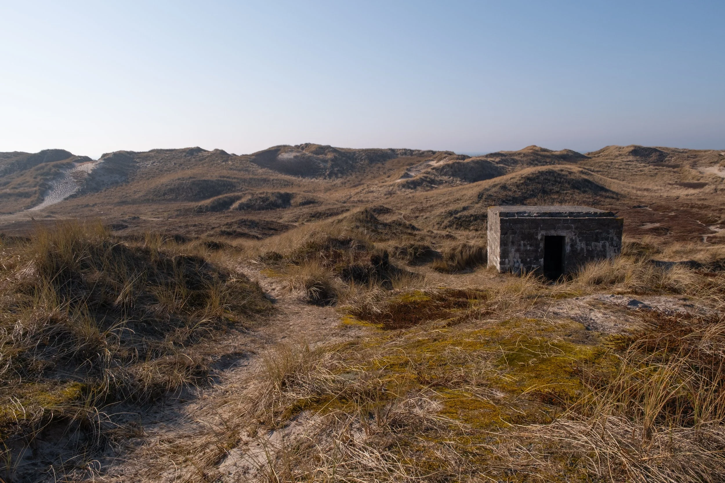 Im rechten Mittelgrund der Fotografie ist ein alter Bunker zu finden, welcher sich in die umliegenden Dünen einbettet. Im Hintergrund sieht man den Weg zwischen den Dünen, welcher zum Strand in Søndervig, Dänemark, führt.