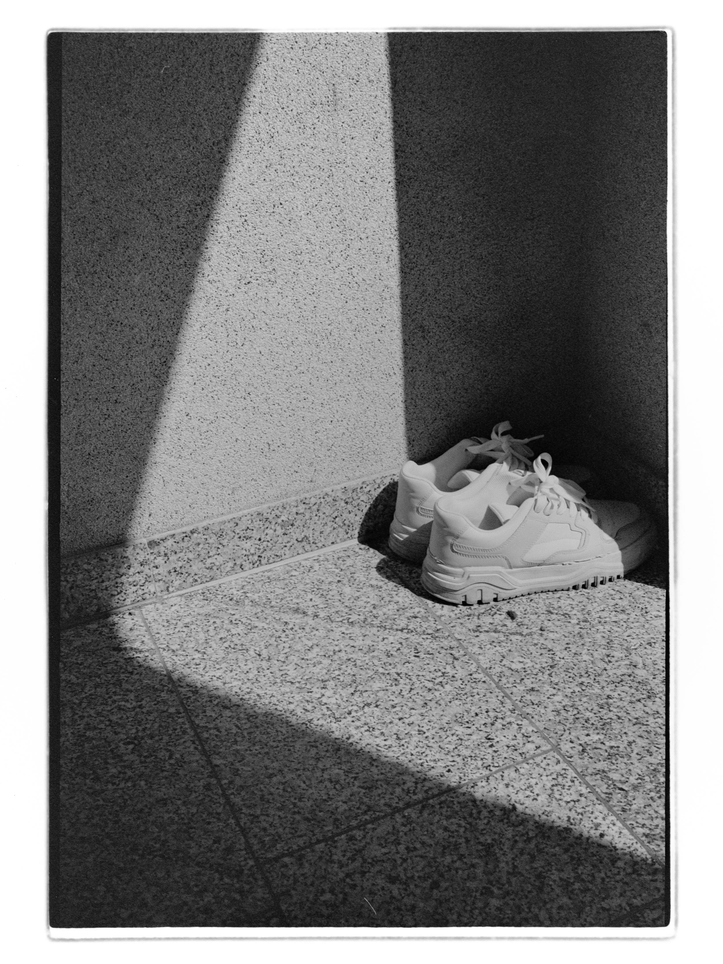 Analoge Fotografie von weißen Turnschuhen auf einem pflegeleichten Steinbodengitter, im Schatten an der Ecke einer Mauer.