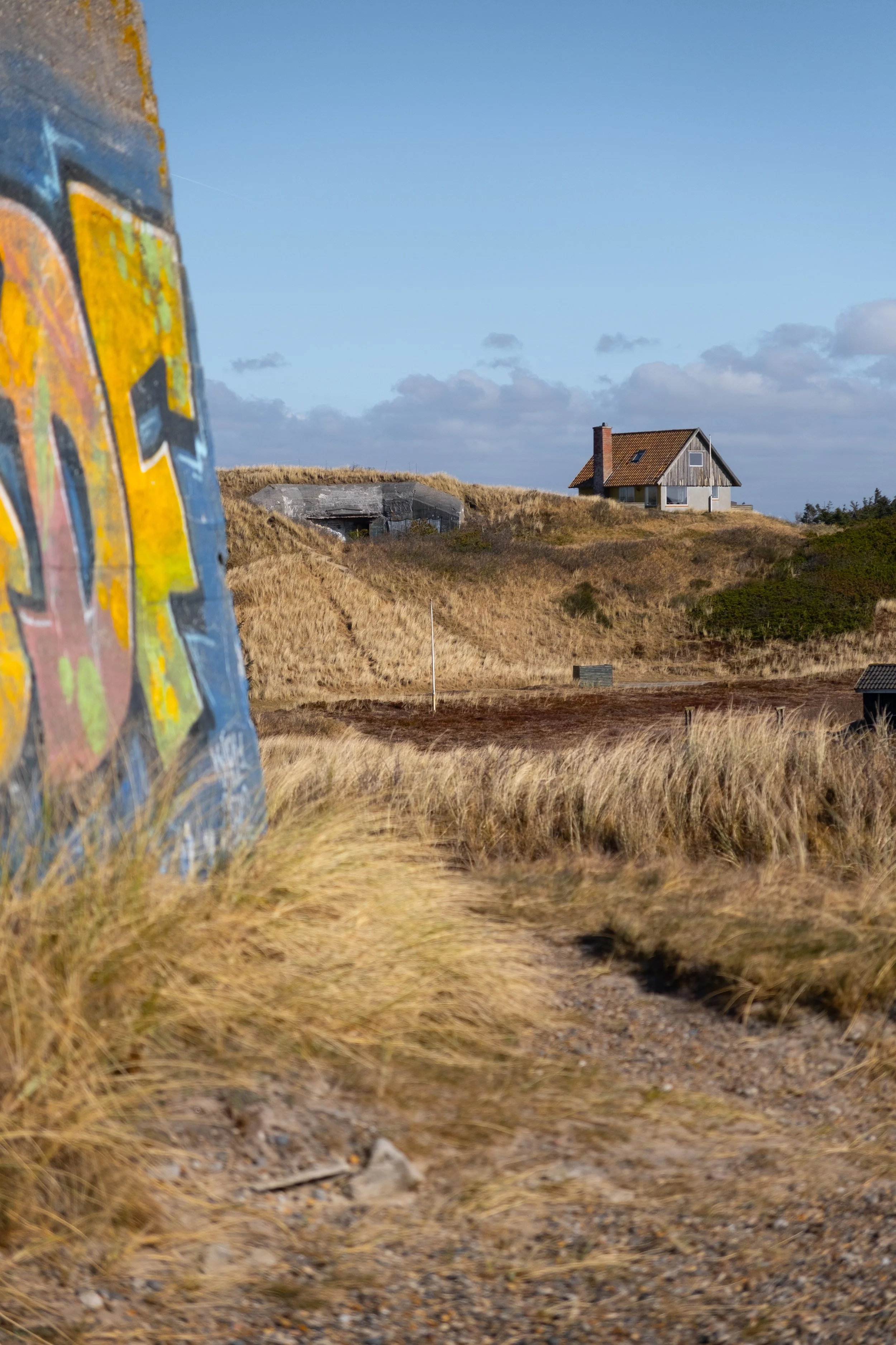 Im Vordergrund dieser Fotografie ist die Wand eines Bunkers zu sehen, welche mit einem Graffiti geschmückt ist. Im Mittelgrund ist das Tal zwischen zwei Dünen zu erkennen. Der Hintergrund zeigt einen weiteren Bunker, neben dem ein Haus steht. Die ges