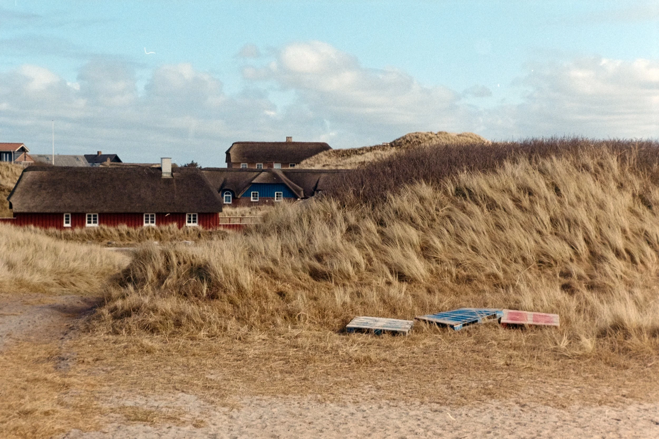 Analoge Fotografie von Häusern in den Dünen von Søndervig in Dänemark. Im Vordergrund befinden sich drei bunte Paletten.