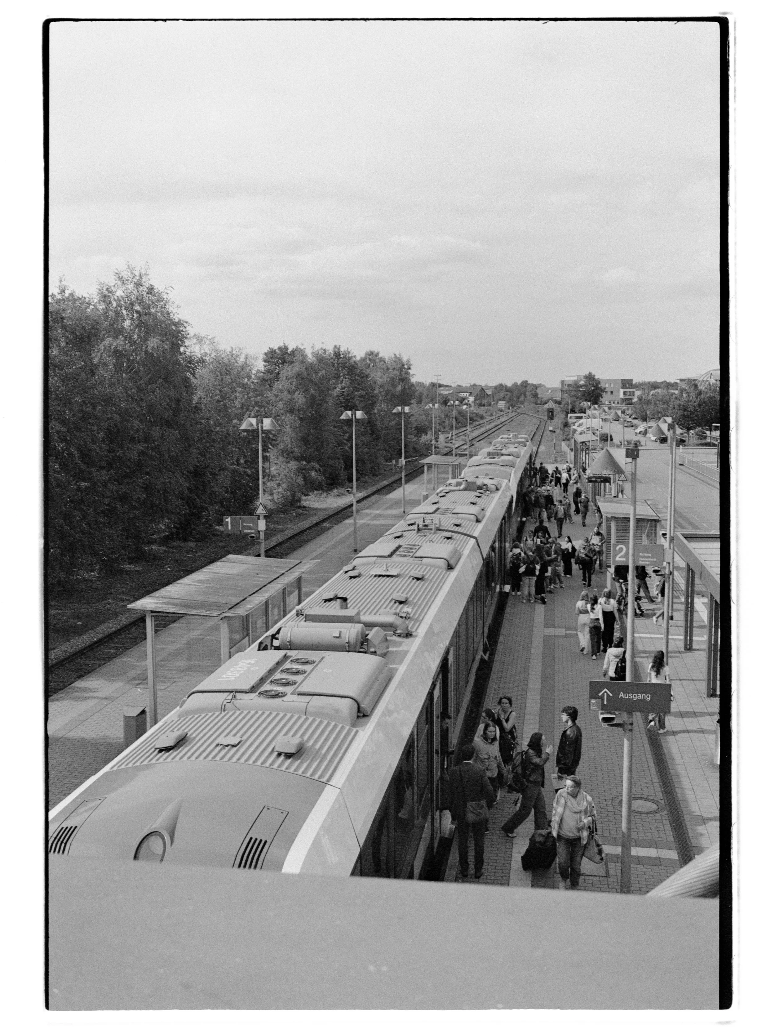 Schwarz-weiß-Foto eines Bahnsteigs in Vechta mit Wartenden und einem Zug, der vor dem Bahnsteig steht. Es sind einige Passagiere mit Gepäck sichtbar. Es gibt Überschriften und eine Beschilderung mit Nummern und einem Ausgangsschild.