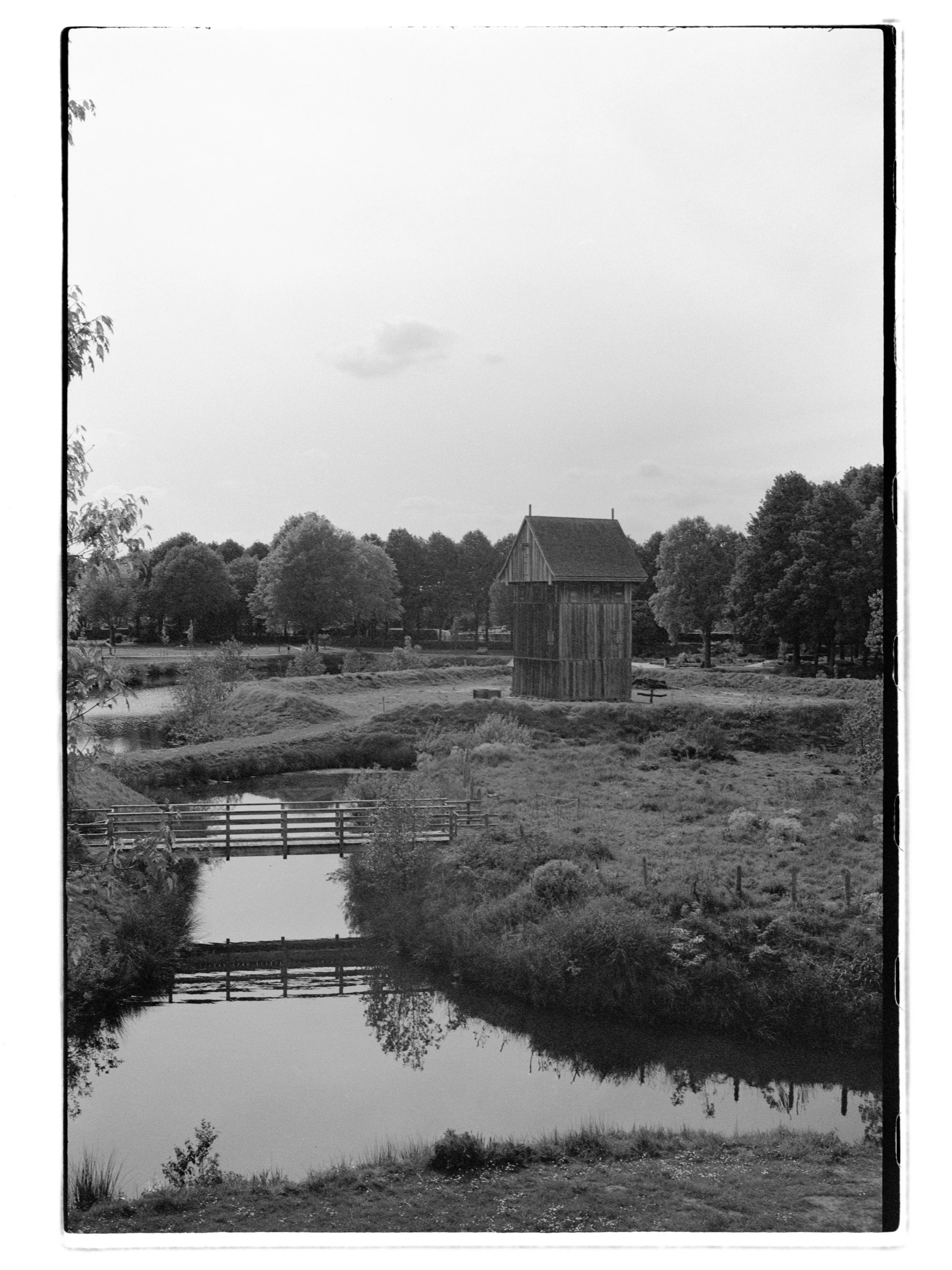Schwarz-weiß-Fotografie eines ländlichen Szenarios mit einem kleinen Fluss, Brücken, Bäumen, und einem großen Holzhaus in der Mitte des Bildes. Das Bild zeigt die Zitadelle in Vechta.