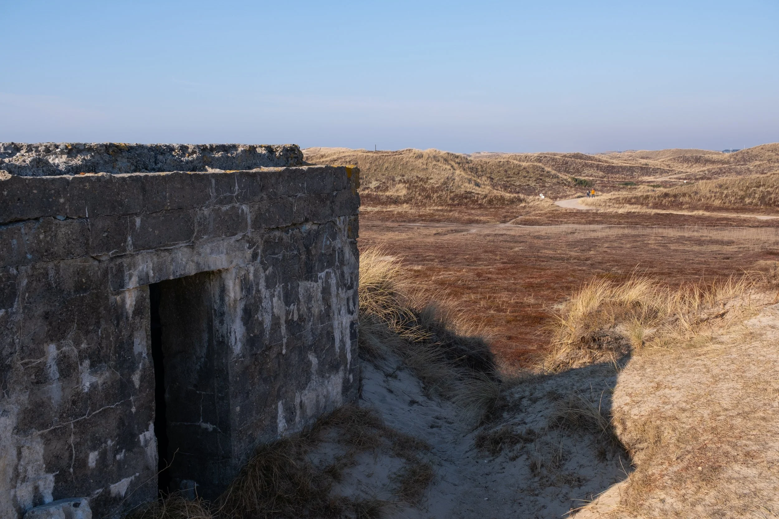 Alter Bunker in einer Dünenlandschaft, umgeben von Sand und trockenem Gras, unter einem klaren blauen Himmel.