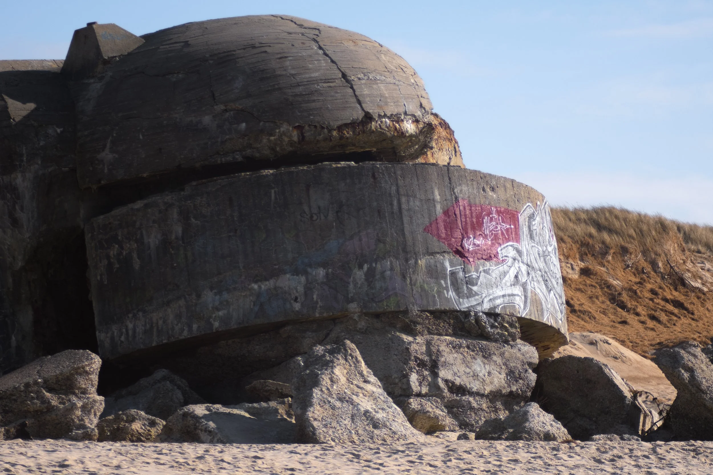 Diese Fotografie zeigt den vorderen Teil eines großen Bunkers in Søndervig. Im Vordergrund ist noch etwas Sand zu sehen, während der Hintergrund von Dünen bedeckt ist.