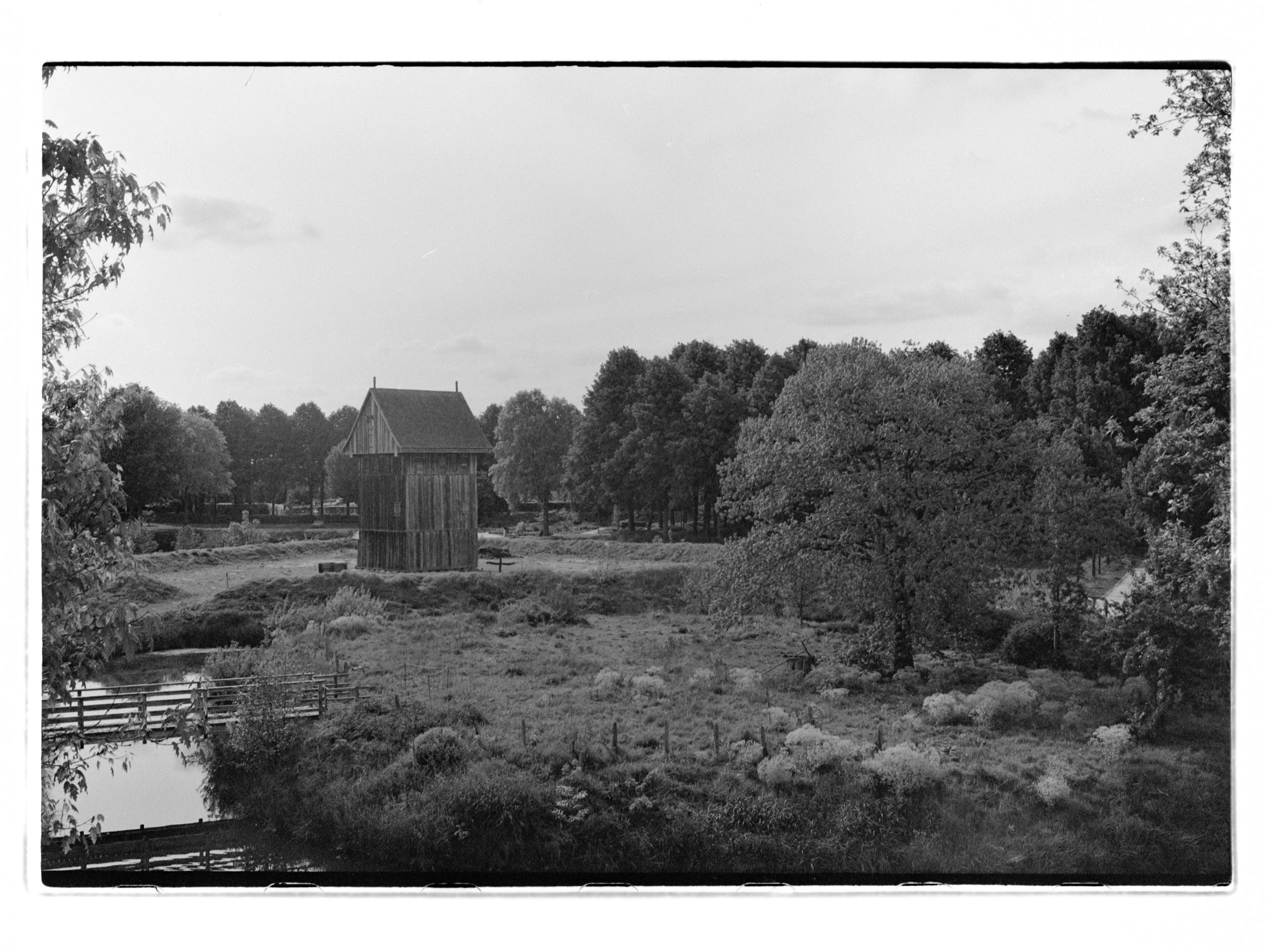 Schwarz-Weiß-Foto eines ländlichen Gartens mit einem alten Holzturm, Bäumen und einem kleinen Teich. Das Bild zeigt die Zitadelle in Vechta.