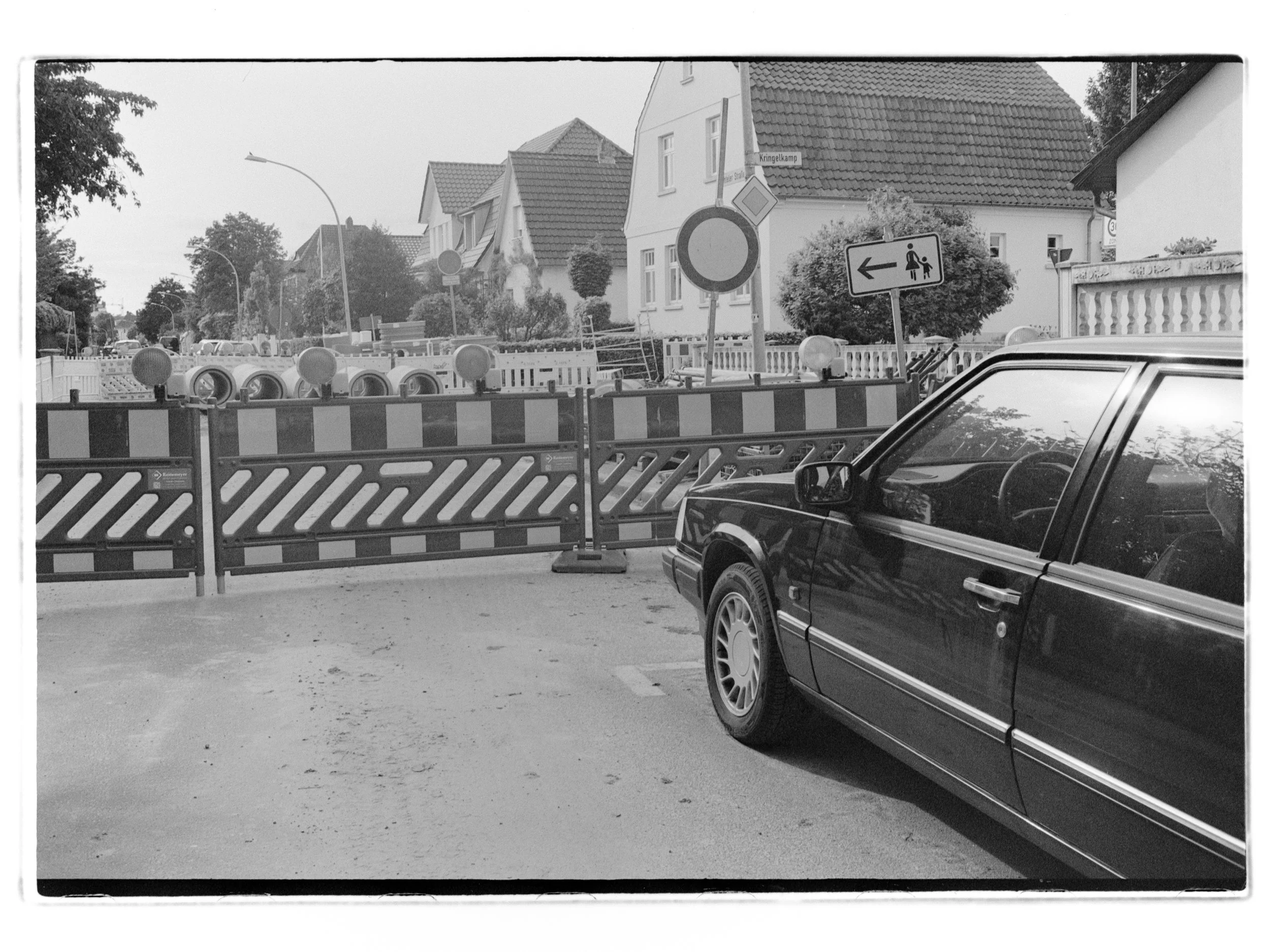 Straßensperrung mit Bauzäunen und Verbotsschild, stehendes Auto in der Nähe, Wohnhäuser im Hintergrund, in Schwarz-Weiß. Das Auto ist ein schwarzer Volvo 740.