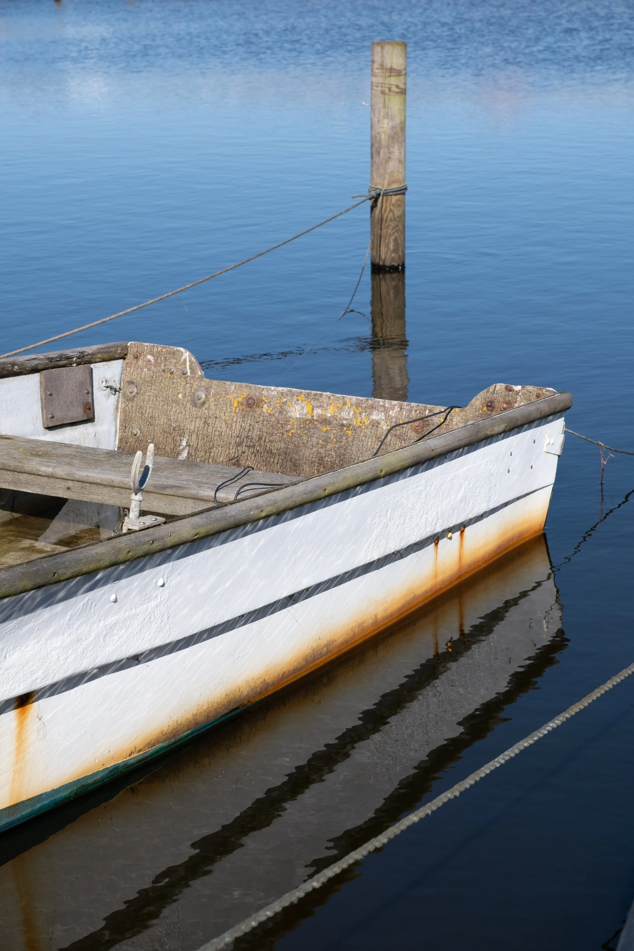 Zu sehen ist ein Abschnitt eines kleinen Ruderboots, welches an einem Holzpfeiler festgebunden ist. Der Rest des Bildes zeigt umliegendes Wasser.