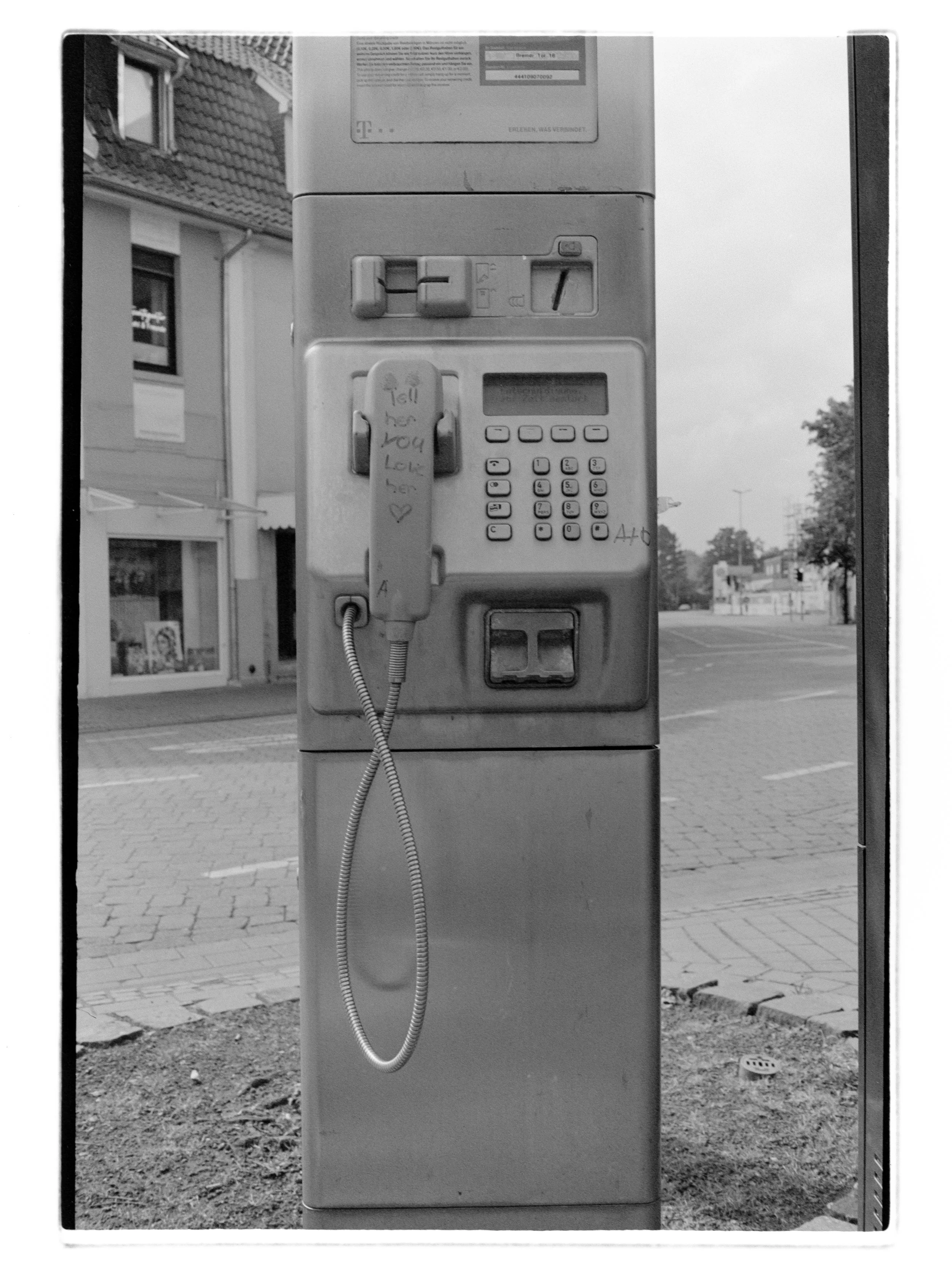 Foto eines öffentlich zugänglichen Telefonautomaten auf einem Bürgersteig, im Hintergrund Gebäude und leeres Straßenbild. Auf dem Telefon steht tell her you love her.