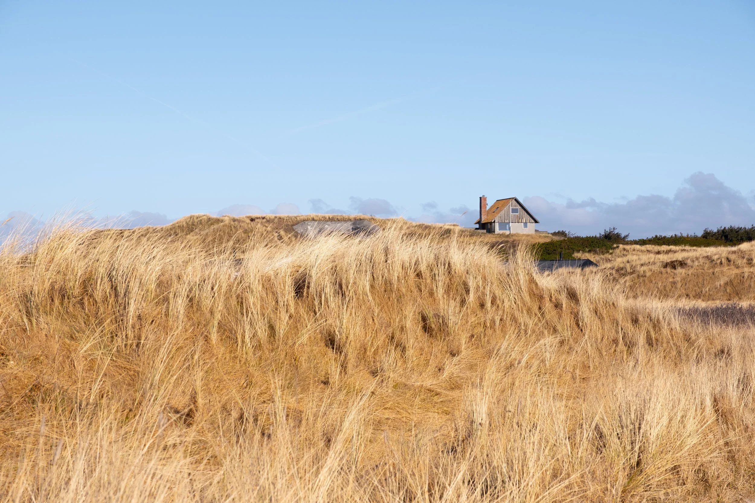 Hinter einer Düne im Vordergrund versteckt sich ein Bunker in der Mitte der Fotografie. Direkt daneben befindet sich ein kleines, einsames Haus auf einer Düne in Søndervig, Dänemark.