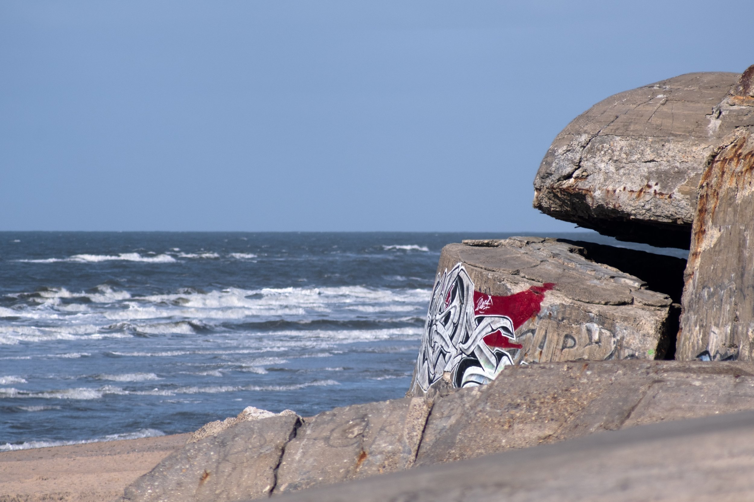 Zu sehen ist ein kleiner Teil eines großen Bunkers am Strand von Søndervig, Dänemark. Im Mittelgrund sieht man die Wällen auf den Strand schlagen, während das Meer im Hintergrund ruhig in den Horizont übergeht.