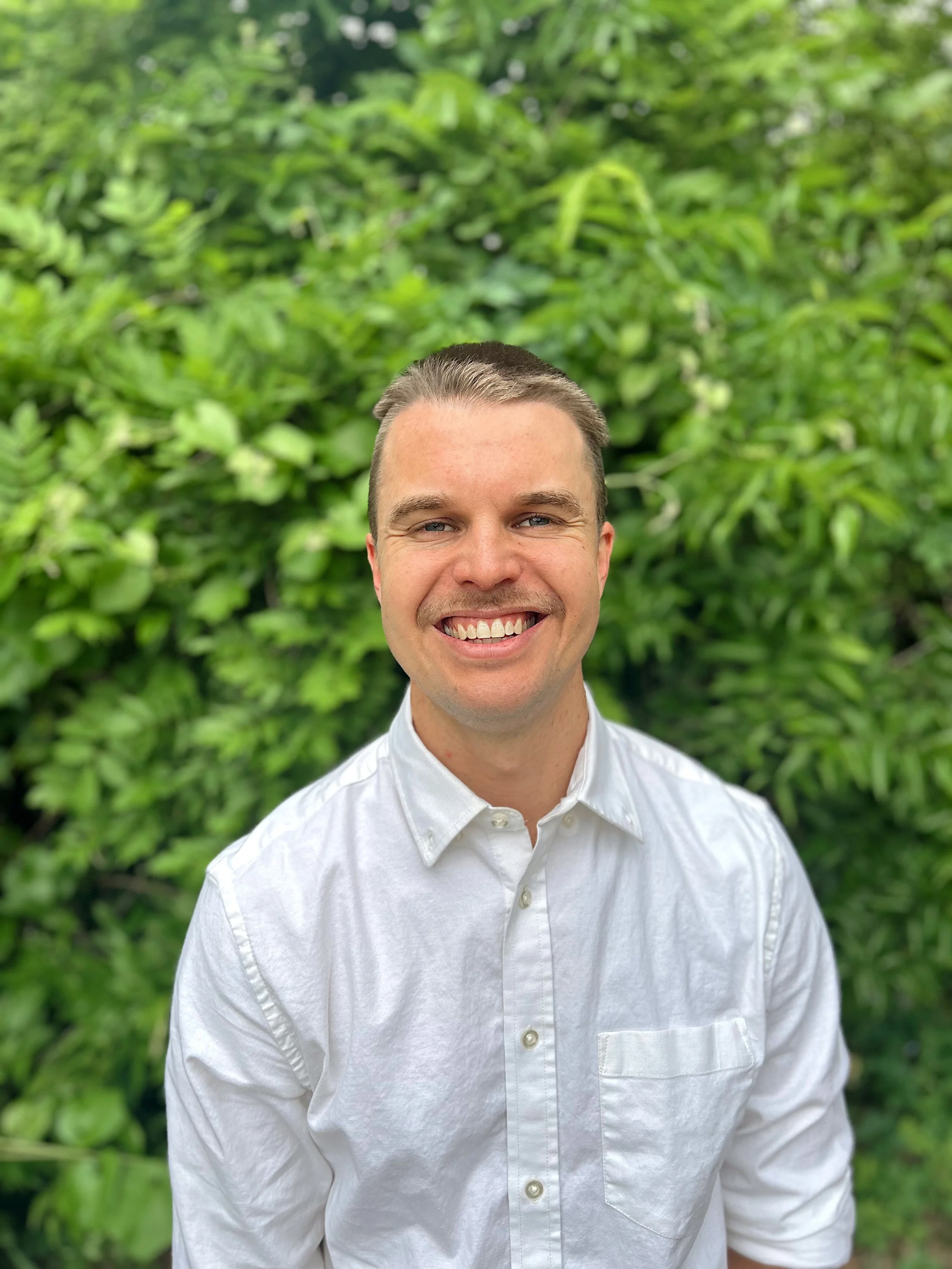 A man smiling outdoors in front of green foliage, wearing a white button-up shirt.