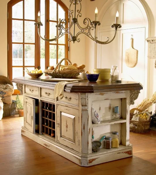 A rustic kitchen island with distressed white paint, topped with bread and bowls. A large window with wooden framing lets in natural light. A chandelier with lit candles hangs above.