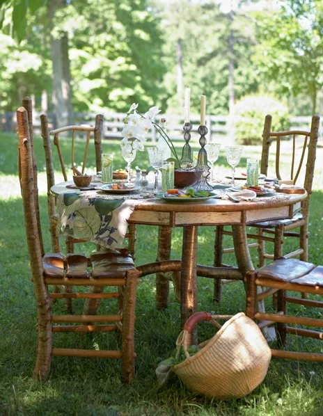 A round outdoor table set for a meal on a grassy lawn, surrounded by wooden chairs with a picnic basket on the ground nearby, with trees and sunlight in the background.