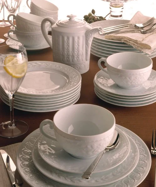 Elegant white dinnerware set with plates, bowls, cups, a teapot, and silverware arranged on a dining table, including a glass of sparkling water with lemon.