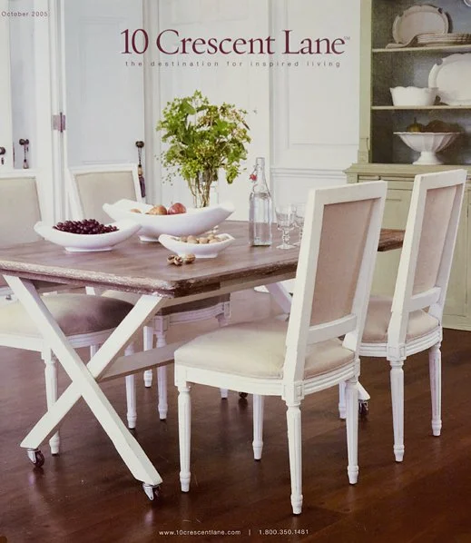 A dining room with a wooden table, four white chairs, a bowl of fruit, a glass bottle of water, and a potted plant. There is a white wall and a green cabinet with dishes.