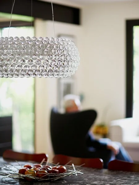 Close-up of a modern hanging chandelier with glass bubble design in a bright living room with a blurred person sitting on a black chair in the background.