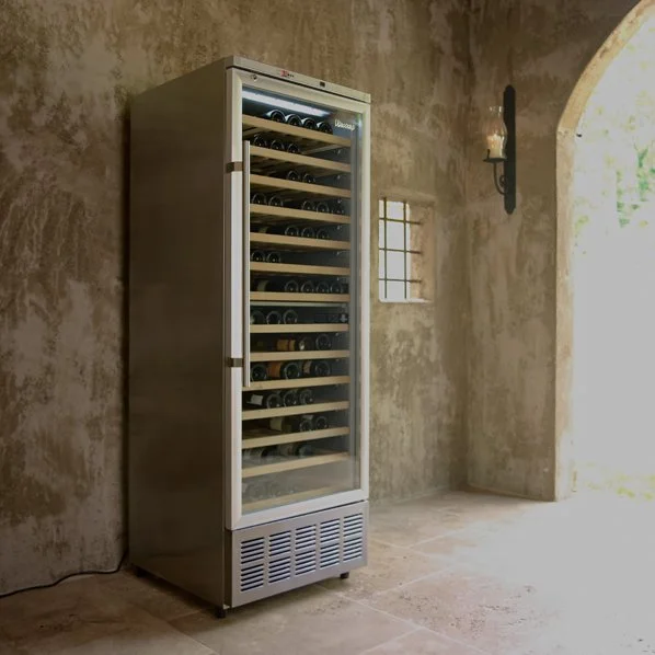 A glass-door wine fridge filled with wine bottles, placed against a rustic stone wall in a room with arched doorway and small window.