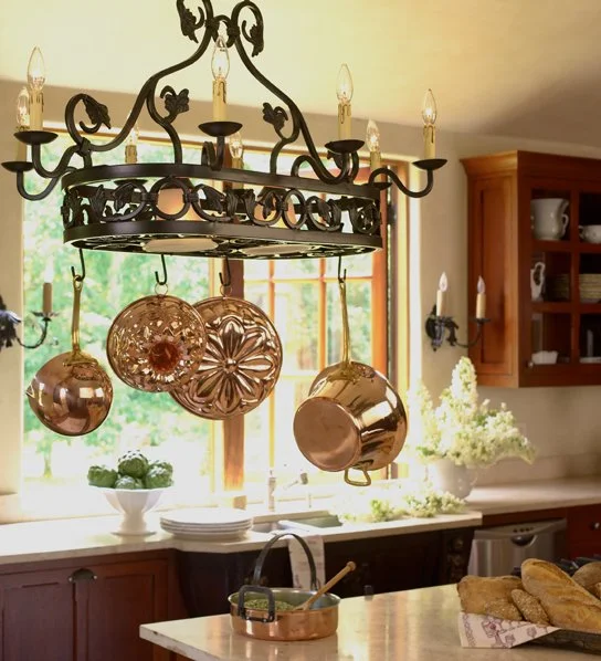 A rustic kitchen with a black wrought iron chandelier holding five candle-shaped light bulbs above a kitchen island. Copper pots and decorative metal plates hang from the chandelier. The background features a large window with a view of greenery, woo