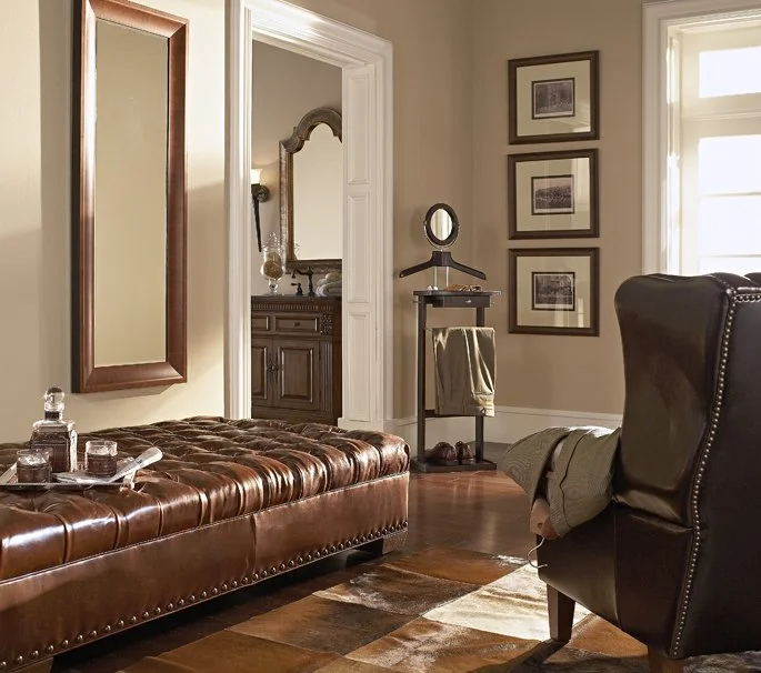 A vintage-style living room with a brown leather tufted ottoman, a black leather armchair, and framed photos on the wall. There is a doorway leading to a bathroom with a wooden vanity and mirror, and a window bringing in natural light.