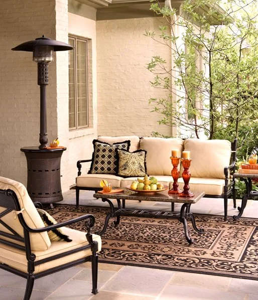Outdoor patio with a black metal and beige cushioned sofa and chairs, a patterned rug, a coffee table with candles and fruit, a tall patio heater, and trees in the background.