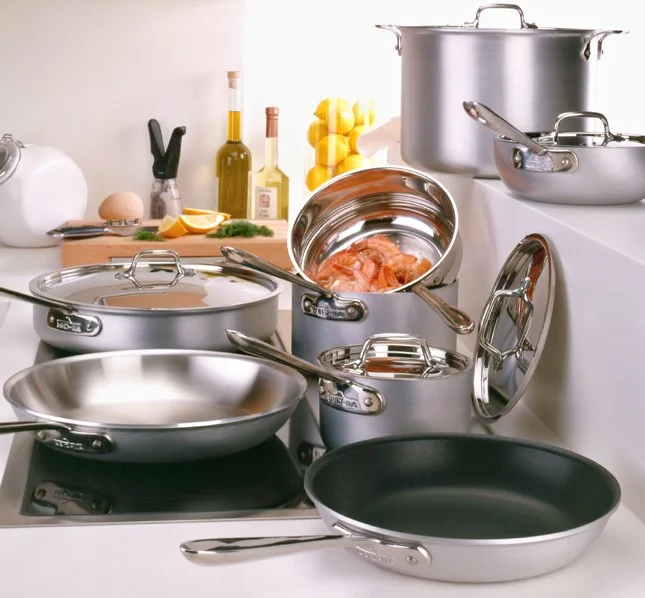 Multiple stainless steel pots, pans, and cookware on a kitchen counter with bottles of oil, lemons, and kitchen utensils in the background.