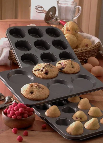 A baking tray with cookie dough in some cups, surrounded by baked cookies, raspberries, eggs, and kitchen utensils on a wooden table.