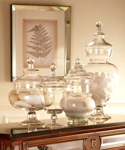 Glass jars with lids filled with cotton balls or similar items on a wooden dresser, with framed botanical print of a fern in the background.