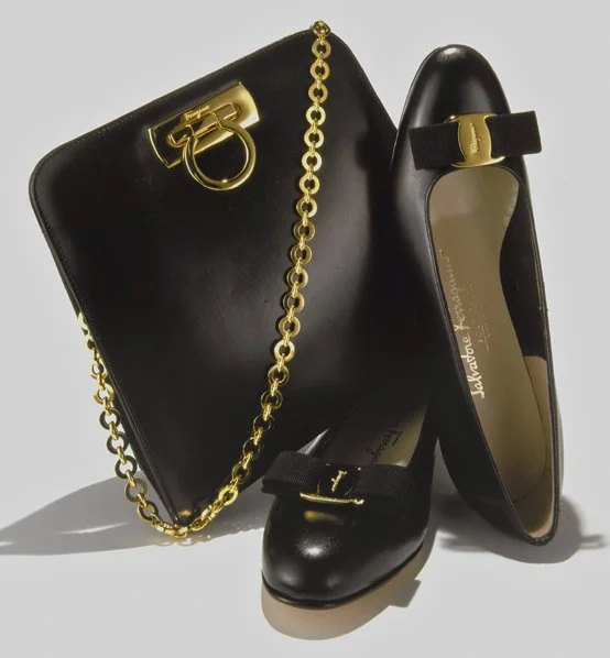 A black designer handbag with a gold chain, gold hardware, and a brand logo, along with black shoes with matching adornments, against a light background.