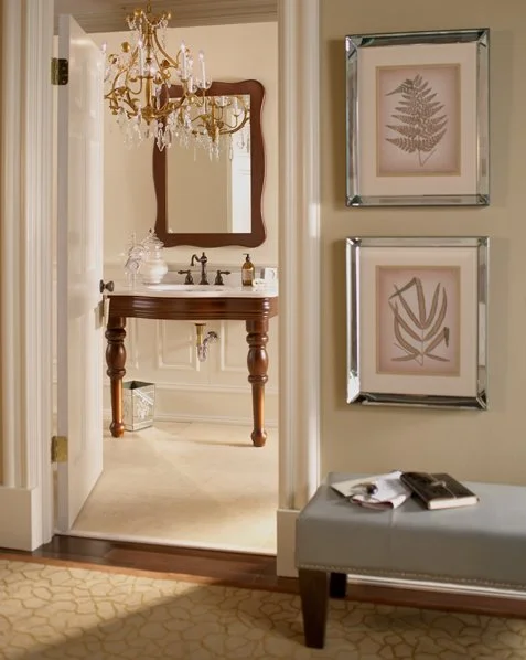 View of an elegant bathroom with a wooden vanity, a mirror, and a chandelier, seen through an open door from a hallway with framed botanical prints and a gray bench.