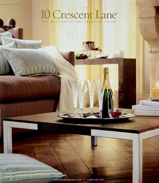 Living room with a brown sofa, pillows, a coffee table with champagne bottle, glasses, and a tray, in front of a window with yellow curtains, with a fireplace. Promotional magazine cover for 10 Crescent Lane.