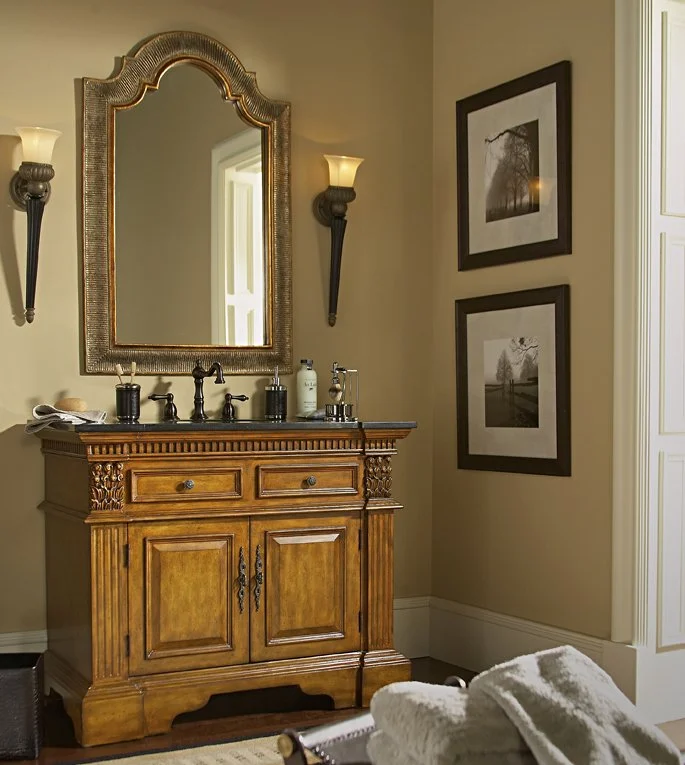 A vintage wooden bathroom vanity with carved details and a dark countertop, topped with various soap and lotion dispensers, reflecting in a large ornate mirror. Two wall sconces with lights frame the mirror, and framed black-and-white photos hang on 