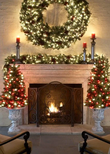A decorated fireplace with a large Christmas wreath hanging above, two small Christmas trees flanking the fireplace, and four red candles on mantelpieces.