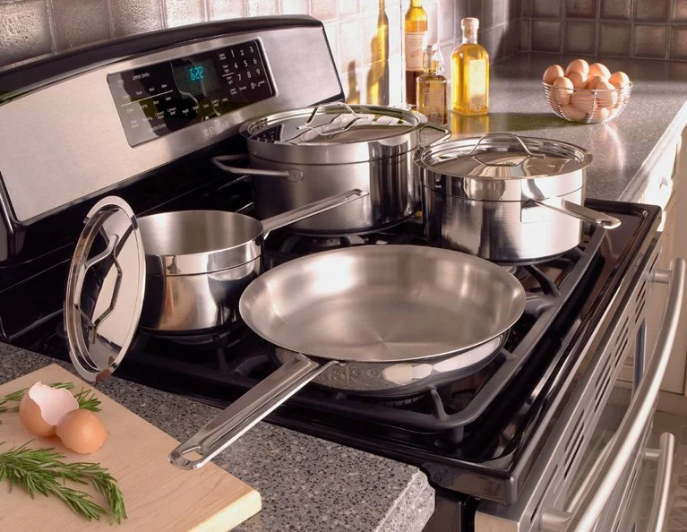 A kitchen stove with multiple stainless steel pots and pans, eggs and greenery on a wooden cutting board nearby, and various bottles on the countertop in the background.