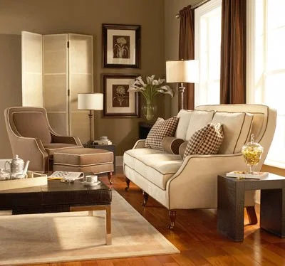 Living room with beige armchair, cream-colored sofa with patterned pillows, side table with yellow flowers, window with brown curtains, and wall art.