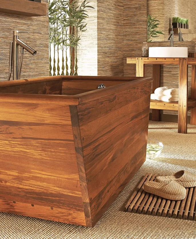 Wooden bathtub in a bathroom with plants, a sink, rolled towels, slippers, and natural decor.