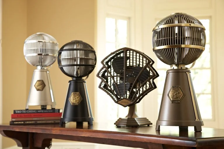 Three vintage-style fans with metal globes on a wooden table, with a decorative fan in the middle, in front of a window with sunlight.