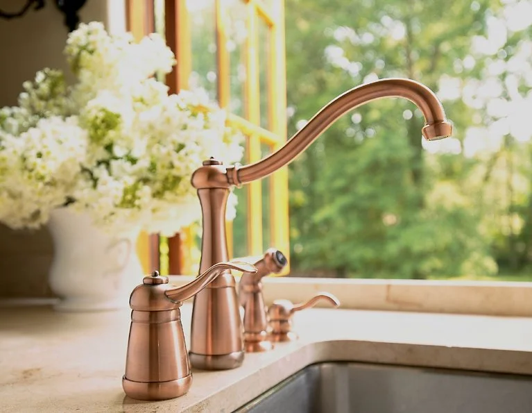 Brass kitchen faucet on a beige countertop with a window showing green trees outside and a white vase with white flowers in the background.