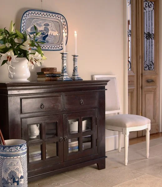 A dark wooden sideboard with glass-paneled doors, a white chair with upholstered seat, and a decorative blue and white plate on the wall. Items on the sideboard include a white vase of white lilies, two blue and white candlesticks with lit candles, a