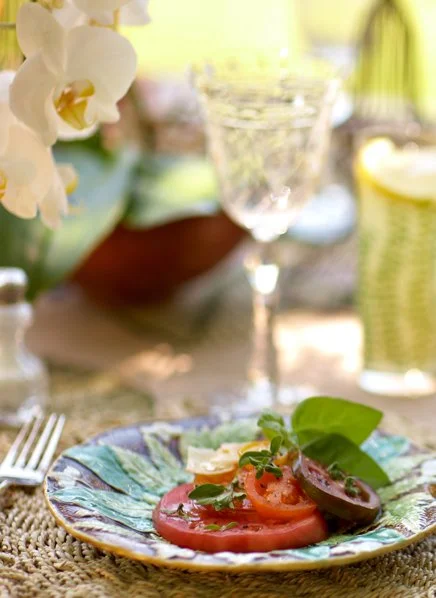 A plate with sliced tomatoes, basil leaves, and cheese on a colorful ceramic dish, with a blurred background of a glass of water, a bowl, and a bottle with lemon in it.