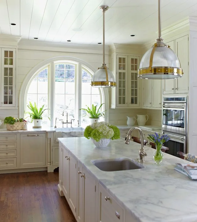 Bright kitchen with white cabinets, a marble island, and a large arched window with sunlight. Green plants are on the windowsill and island, with hardwood floors and modern pendant lights above the island.