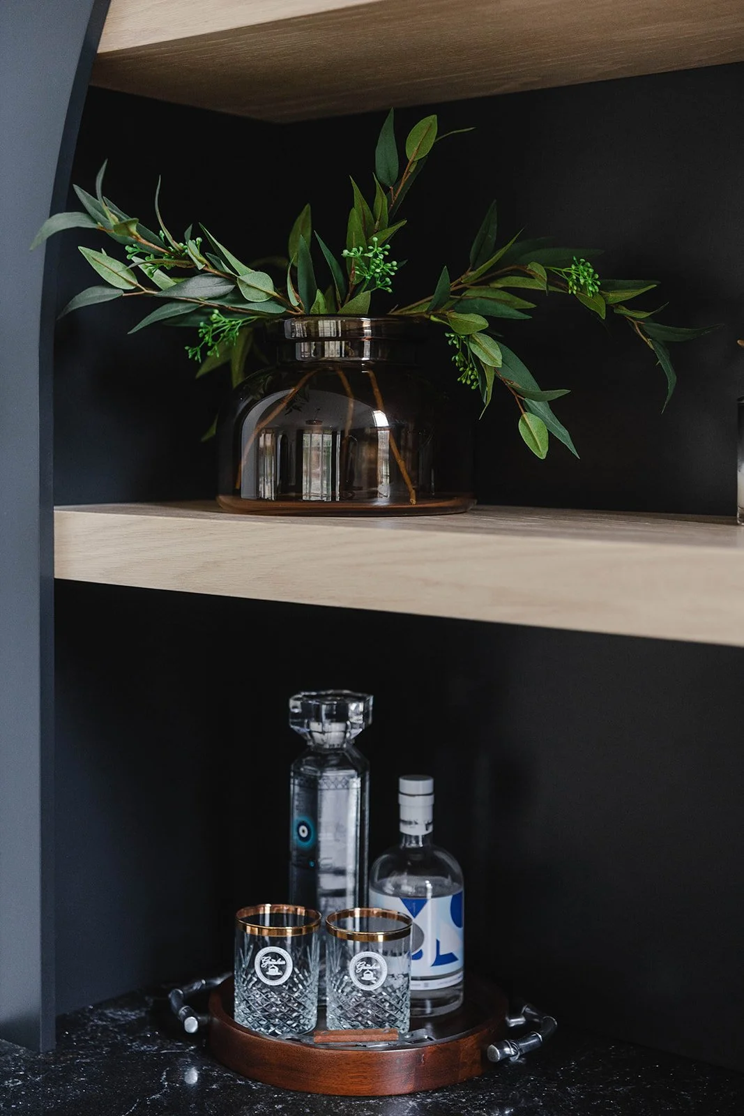 A black and wooden shelf with a dark glass vase containing green leafy branches on the top shelf, and a round wooden tray holding two bottles of alcohol with two crystal glasses with gold rims in front on the bottom shelf.