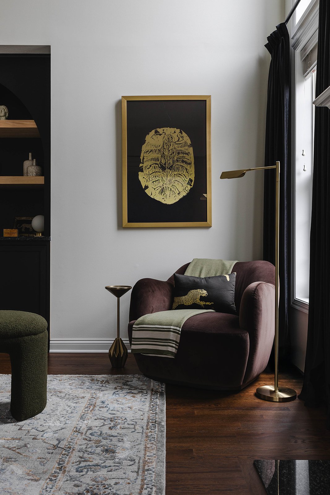 Living room corner with brown armchair, black and gold lamps, framed art on wall, with black curtains and a window, and a patterned rug on wooden floor.