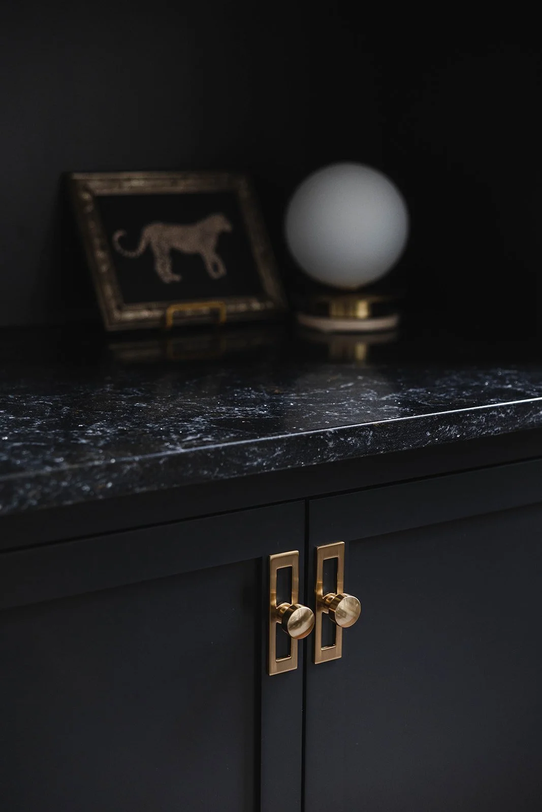 Black cabinet with gold handles, black marble countertop, decorative framed lion artwork, and a round white lamp or globe on a black base.