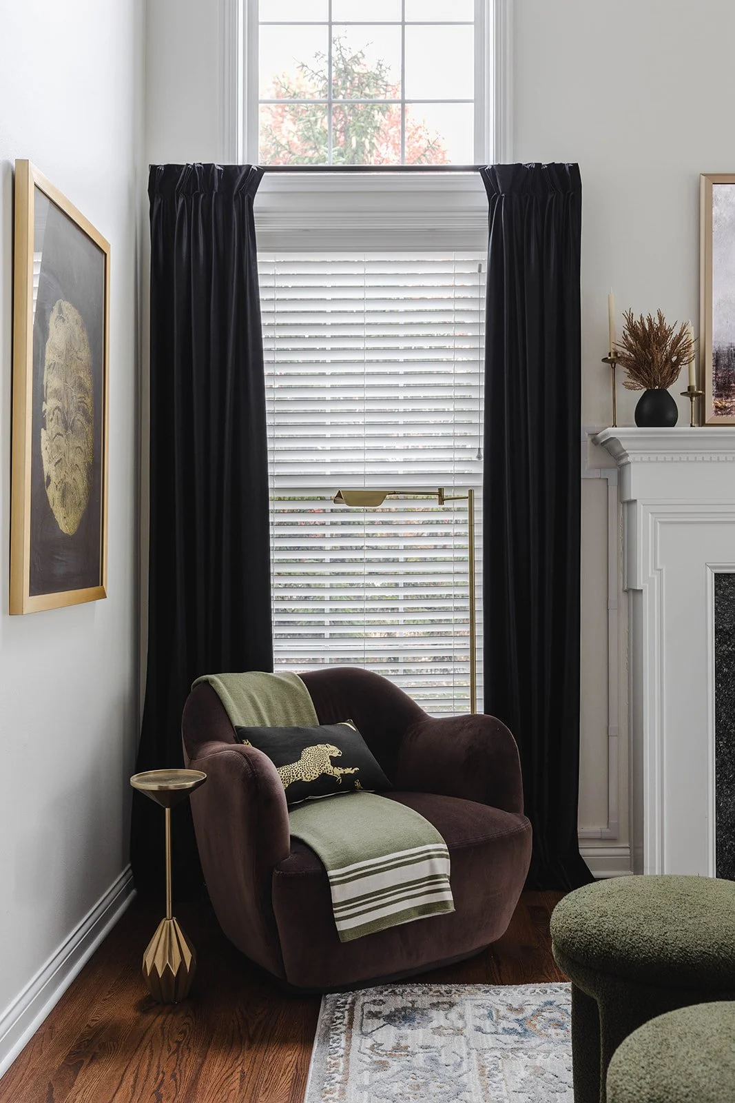 A cozy living room corner with a brown velvet armchair, a green throw blanket and cushion, a gold side table, black curtains on a large window, and a white fireplace with decor.