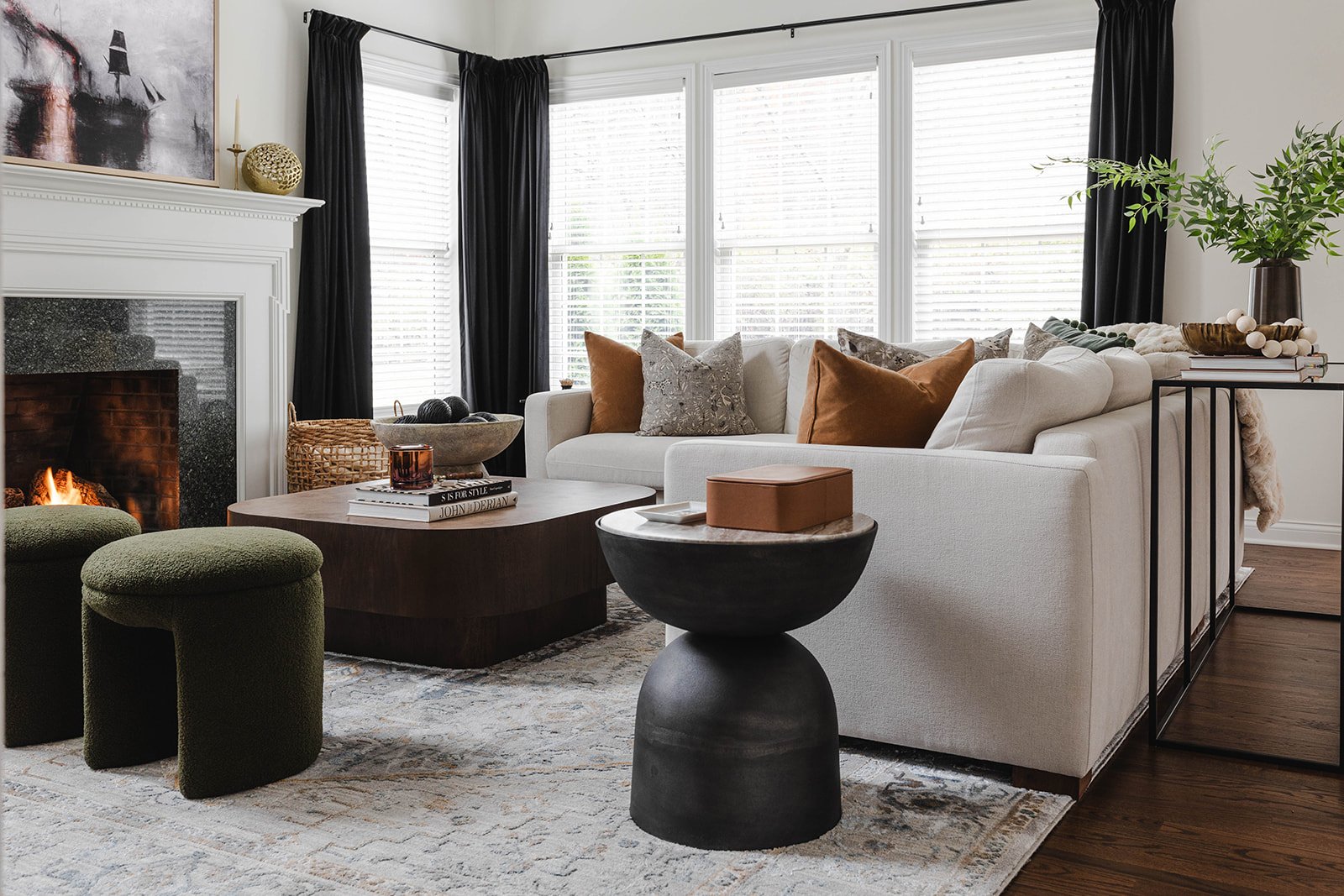 Living room with a white sofa, colorful pillows, a fireplace, a wooden coffee table, two green ottomans, and a side table with decor, illuminated by natural light through large windows with black curtains.